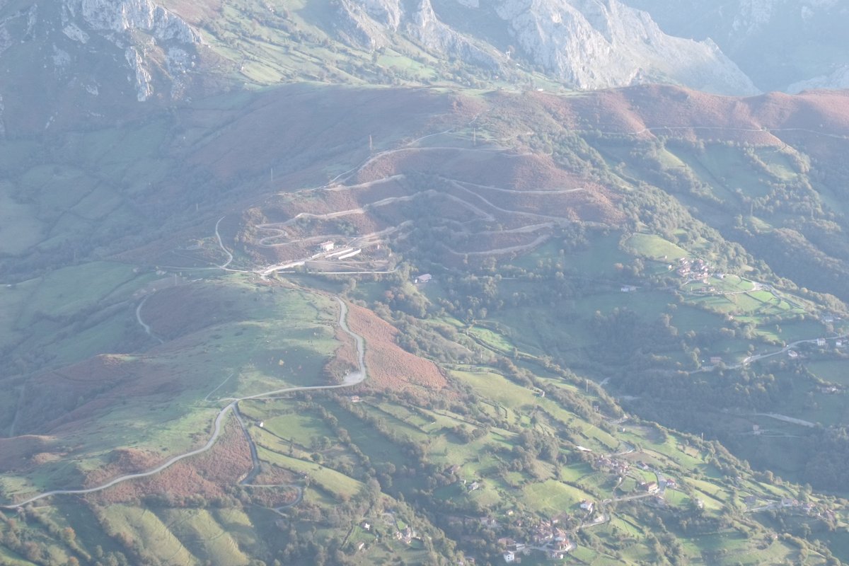 Looking down from the Angliru road on the Viapará picnic area and the zigzagging mountain road above La Vega de Riosa, with green meadows and the limestone cliffs of the Sierra del Aramo