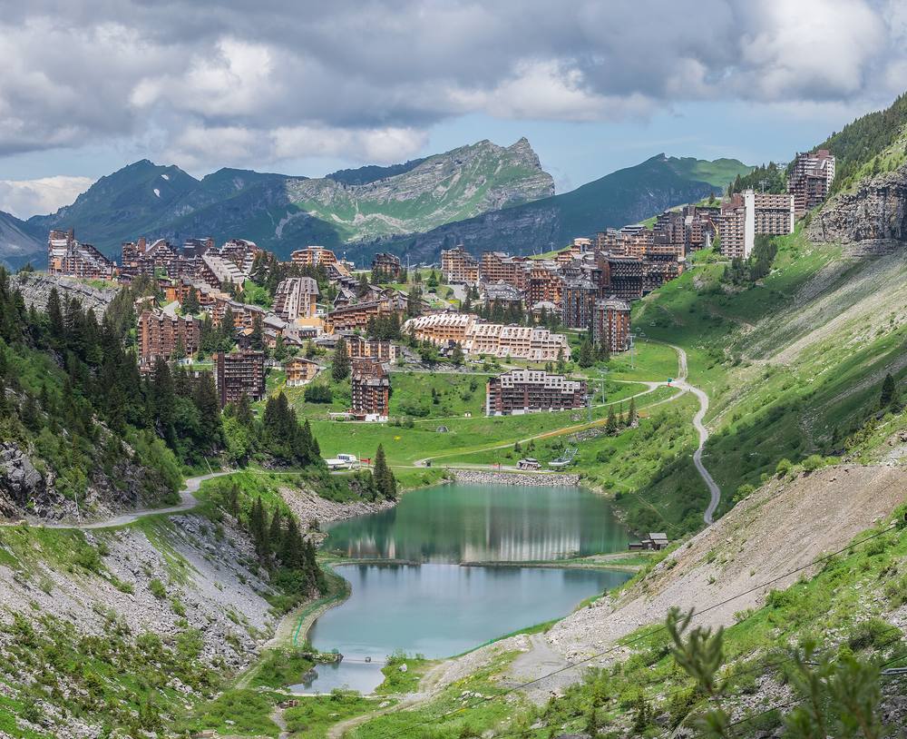 Summer view of Avoriaz: the wood-clad resort buildings perched on cliffs above a turquoise lake, with a winding road, green slopes and rocky Chablais peaks behind