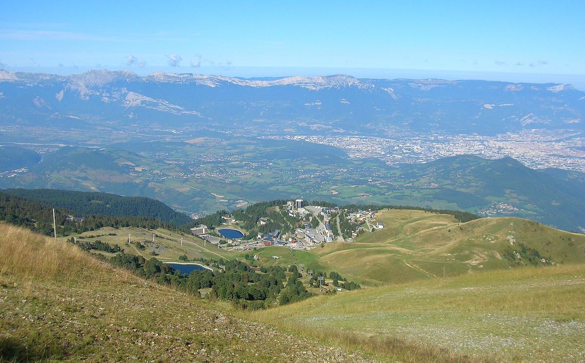 Summer view from the slopes of Chamrousse over the Le Recoin resort and forest down to the Grenoble valley, with distant mountain ranges on the horizon