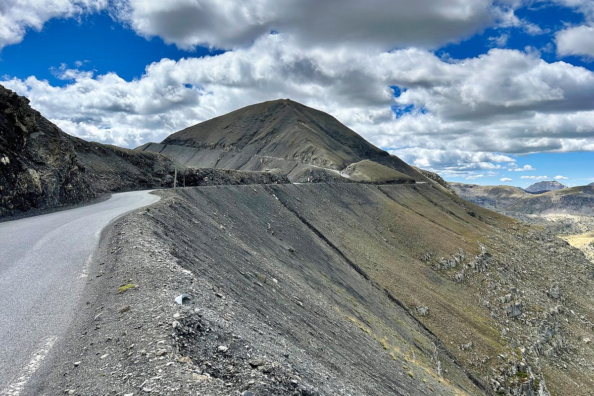 The road near the Col de la Bonette running along a bare grey scree slope, with the cone of the Cime de la Bonette and the loop road cut across it under a cloudy blue sky