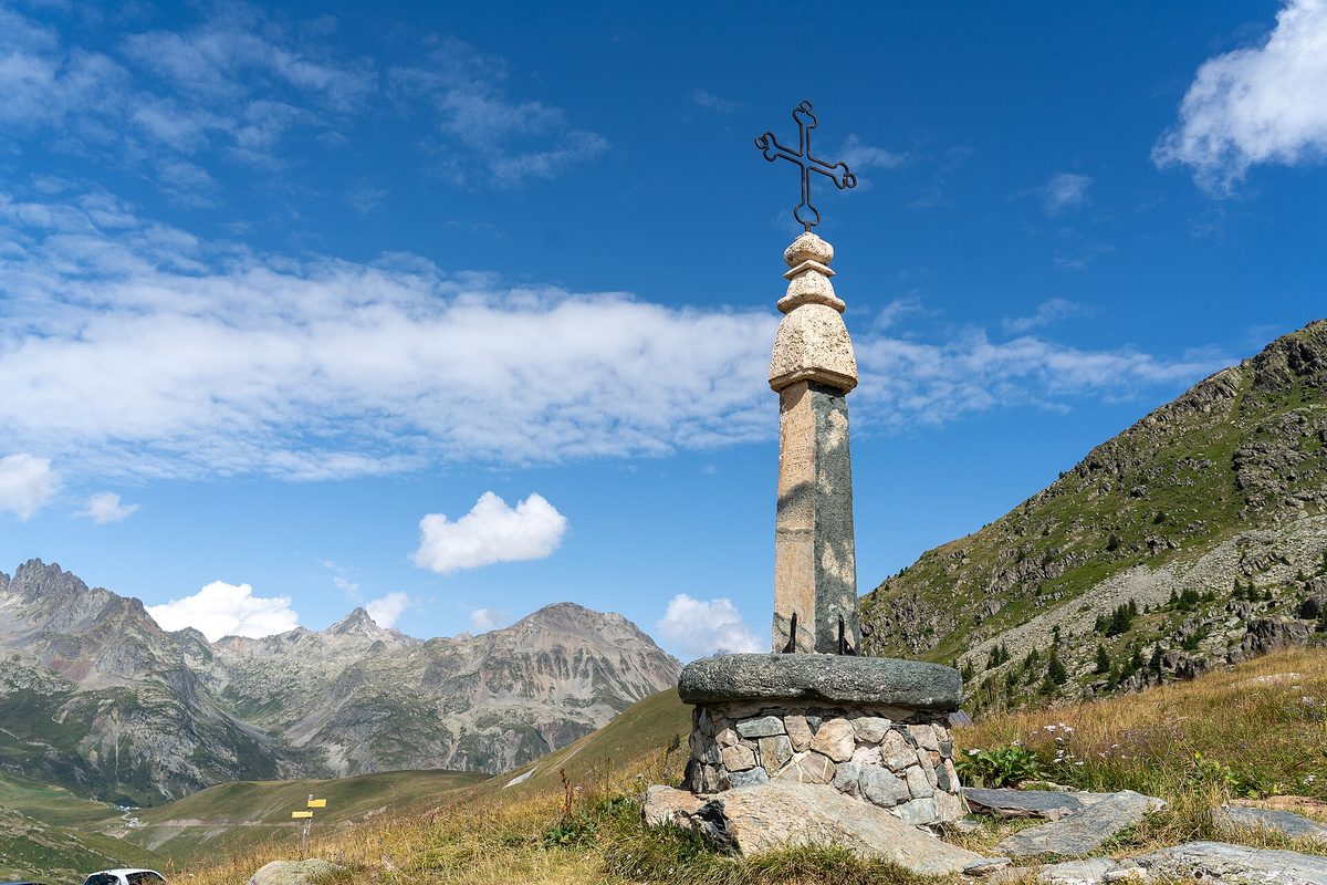 The iron cross on its stone base at the Col de la Croix de Fer, with mountains and a blue sky behind