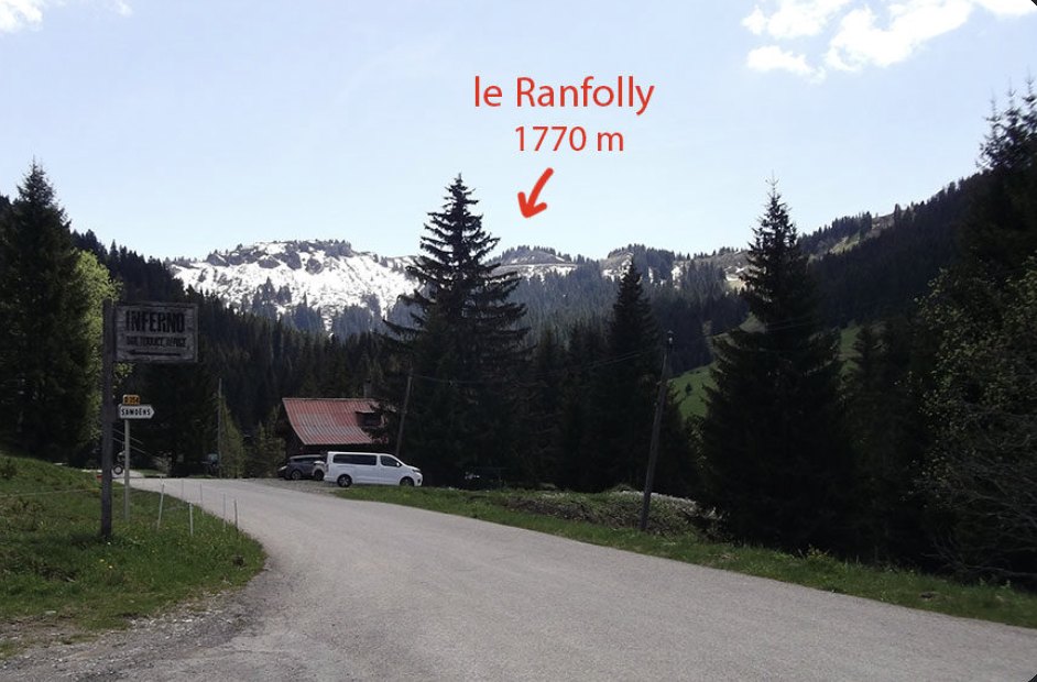 Quiet road near Les Gets with an Inferno signpost, a chalet and pine trees, and the snow-streaked Ranfolly summit marked 1770 m in red above