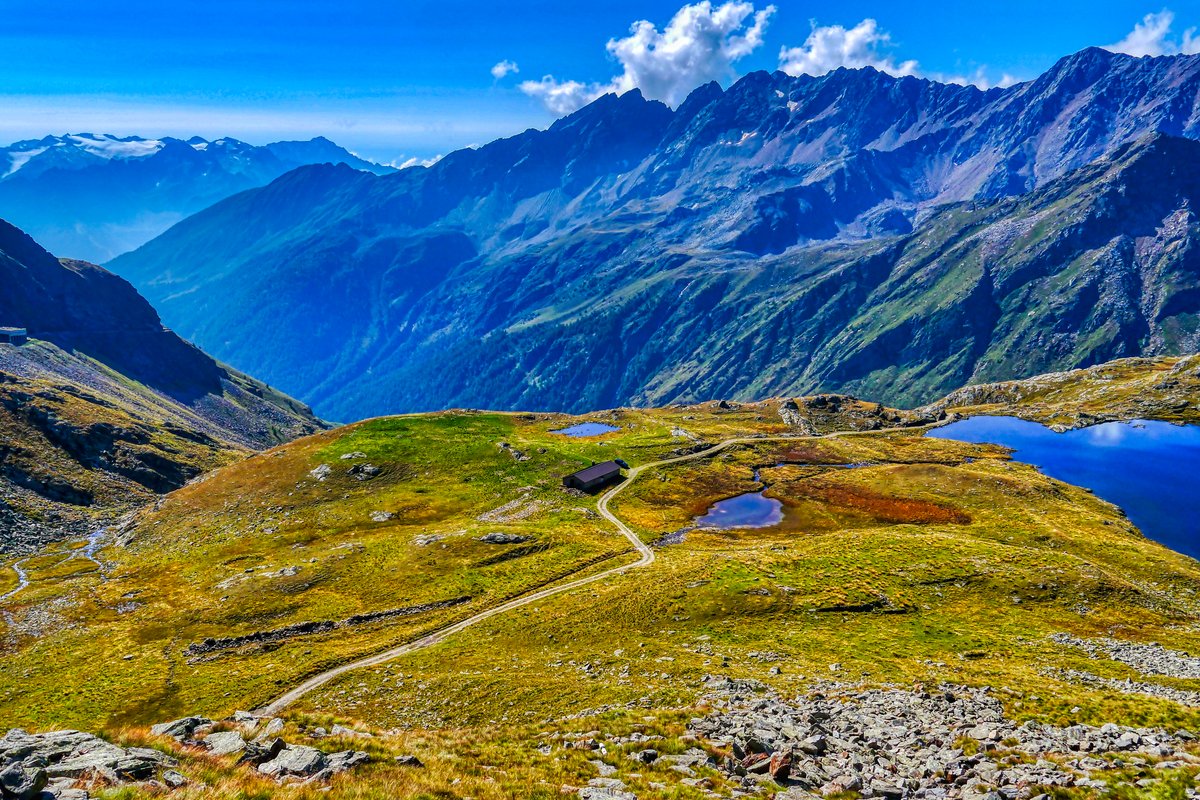 The south side of the Passo di Gavia: a narrow road winding across a high grassy plateau with small lakes, below a long ridge of dark mountains under a blue sky