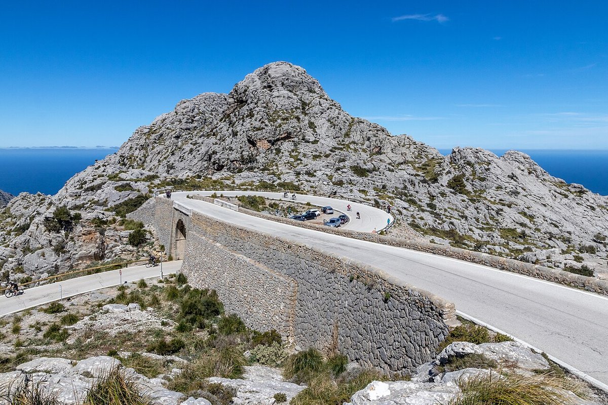 The Nus de sa Corbata on the road to Sa Calobra: the road curves over a stone bridge and loops back underneath itself below a grey limestone peak, with cyclists on the bend and the sea behind