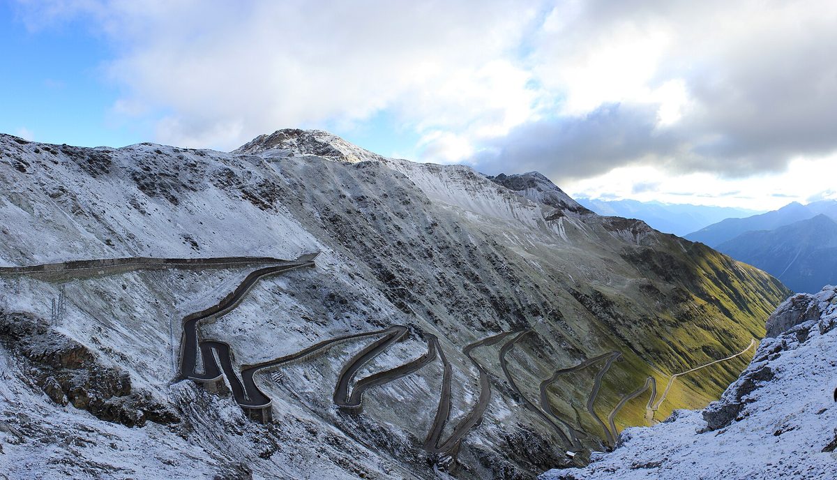 The north side hairpins of the Passo dello Stelvio zigzagging up a snow-dusted mountainside under a cloudy sky