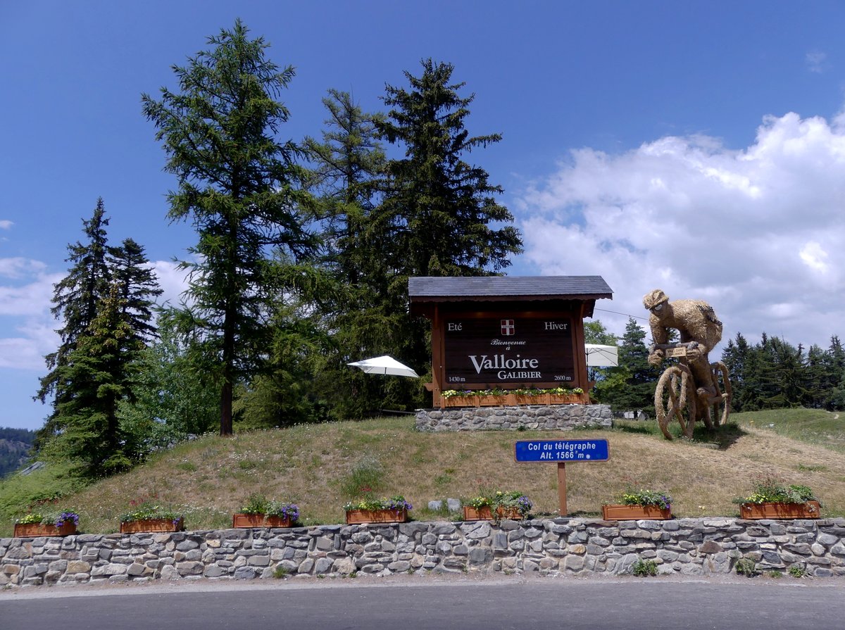 Summit of the Col du Télégraphe at 1,566 m: the wooden Valloire Galibier welcome sign, the blue pass sign, and a large straw cyclist sculpture under a blue sky